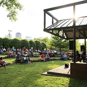a group of people sitting down on a lawn listening to live music in a park.